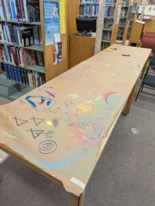 A long library table covered with brown paper shows colorful chalk drawings and scribbles, with markers and crayons scattered on top. Bookshelves are visible in the background.