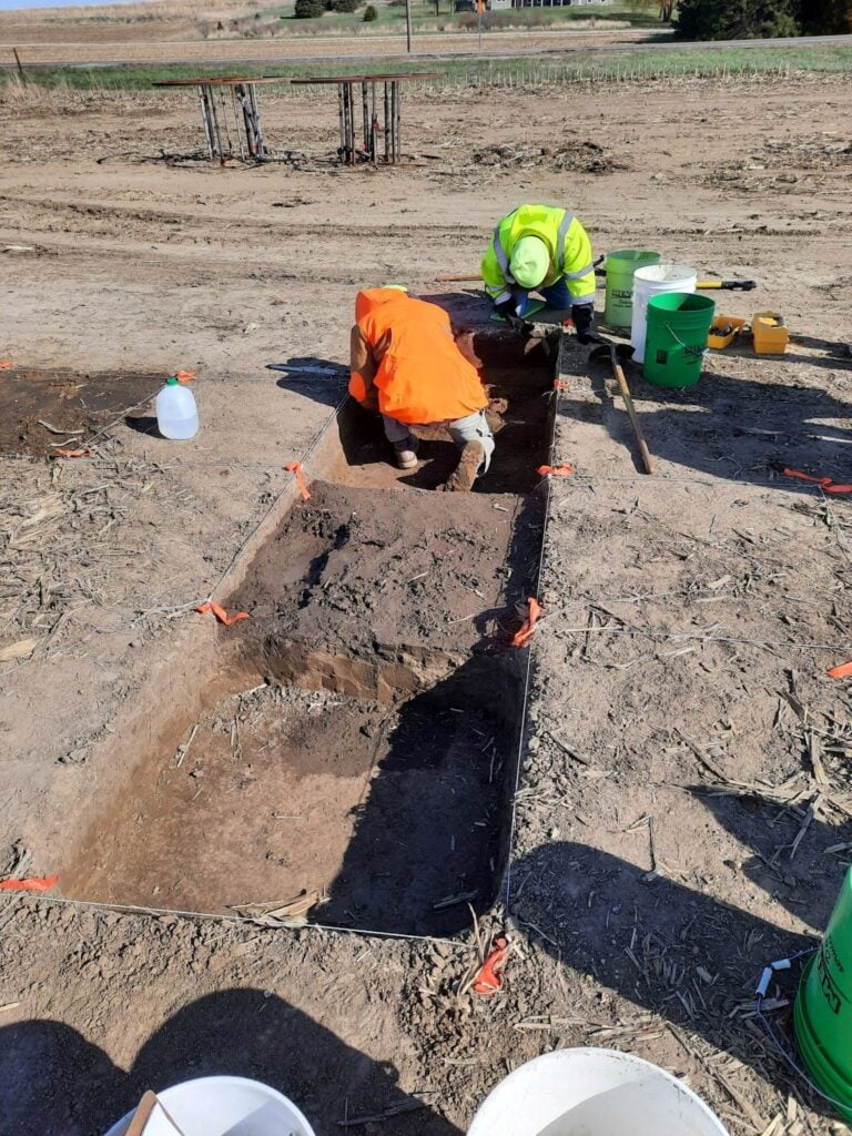 Two people in safety vests are excavating a rectangular trench in a dry, open area; tools and buckets are visible nearby.