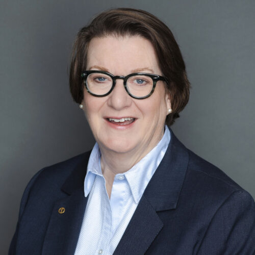 A person with short brown hair, wearing glasses, a navy blazer, and a light blue shirt, smiling in front of a plain gray background.