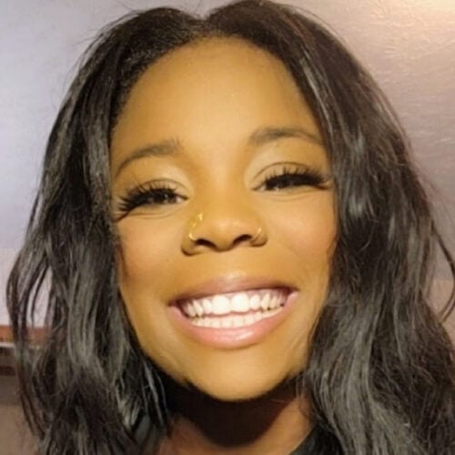 A woman with long black hair smiles at the camera, showing her teeth. She has two nose rings and is indoors with a light-colored wall in the background.