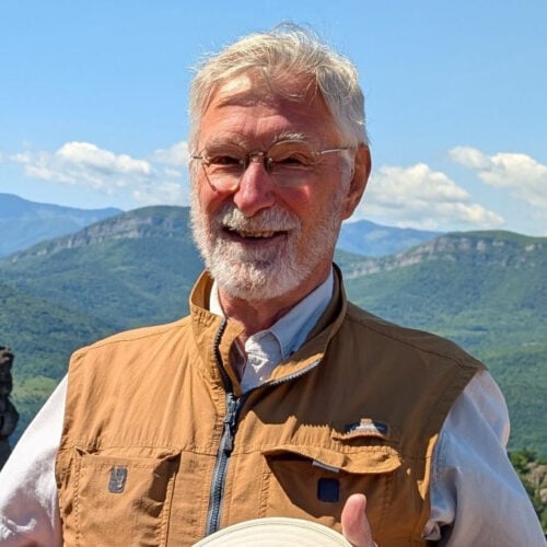 An older man with gray hair and a beard, wearing glasses and a tan vest, smiles outdoors with mountains and a blue sky in the background.