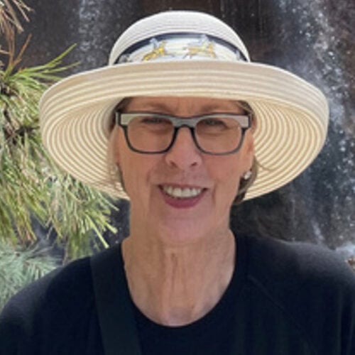A person wearing glasses and a wide-brimmed hat smiles at the camera outdoors, with greenery and a dark background behind them.