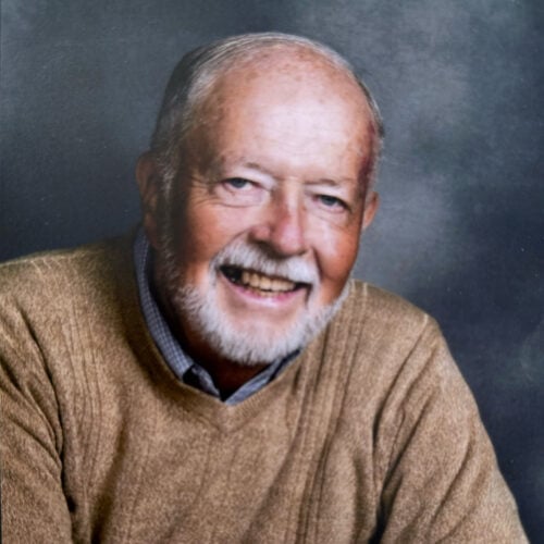 An elderly man with a white beard and hair, wearing a tan sweater over a checkered shirt, smiles at the camera against a dark, neutral background.