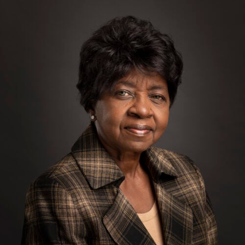 Portrait of an older woman with short curly hair, wearing a brown plaid blazer and beige top, posing in front of a plain dark background.