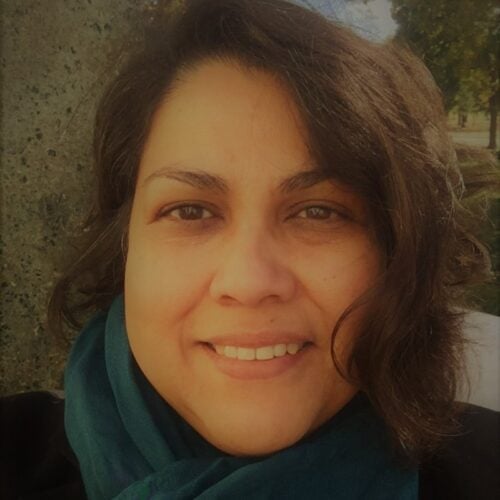 A person with medium-length brown hair smiles at the camera, wearing a teal scarf and dark top, outdoors with trees and a stone surface in the background.