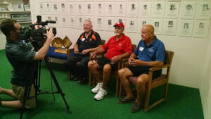 A cameraman films three older men sitting on chairs, giving an interview in front of a wall covered with plaques or certificates.