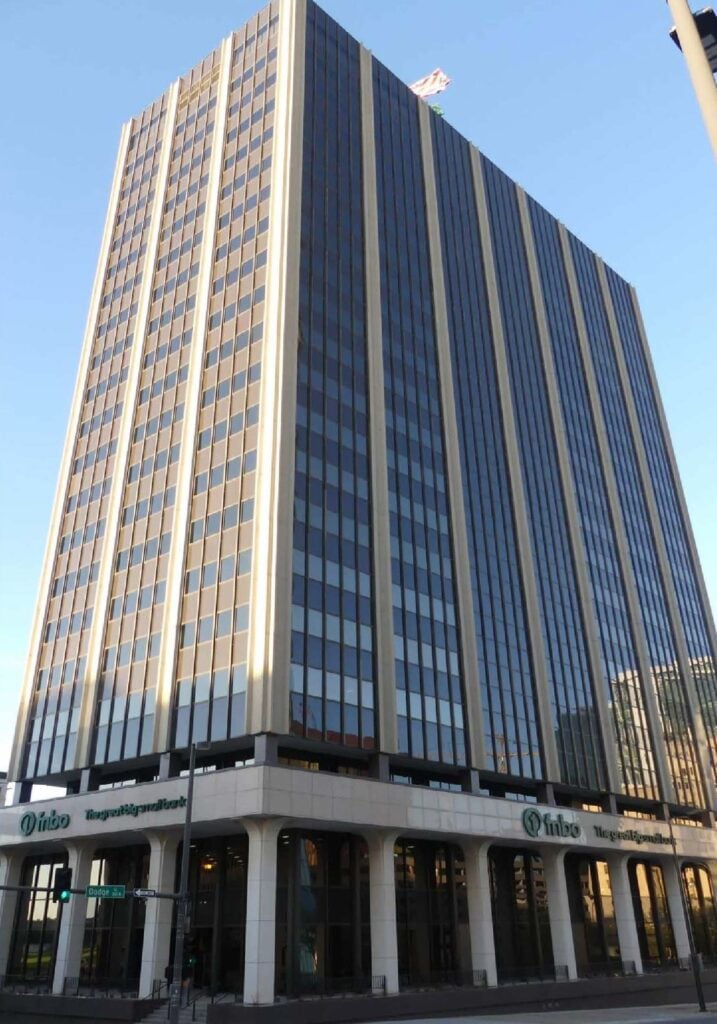 Tall office building with glass windows and vertical stripes, featuring a bank's signage at street level and a flag on the roof, viewed from a low angle.