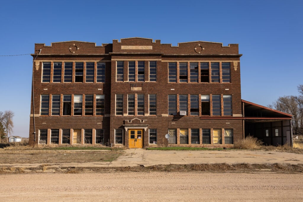 A three-story abandoned brick high school building with many broken windows and boarded-up doors, under a clear blue sky.