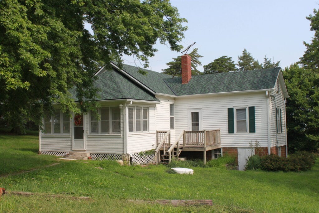 A white single-story house with green roof shingles, a brick chimney, a screened porch, and a small wooden deck, surrounded by grass and trees.