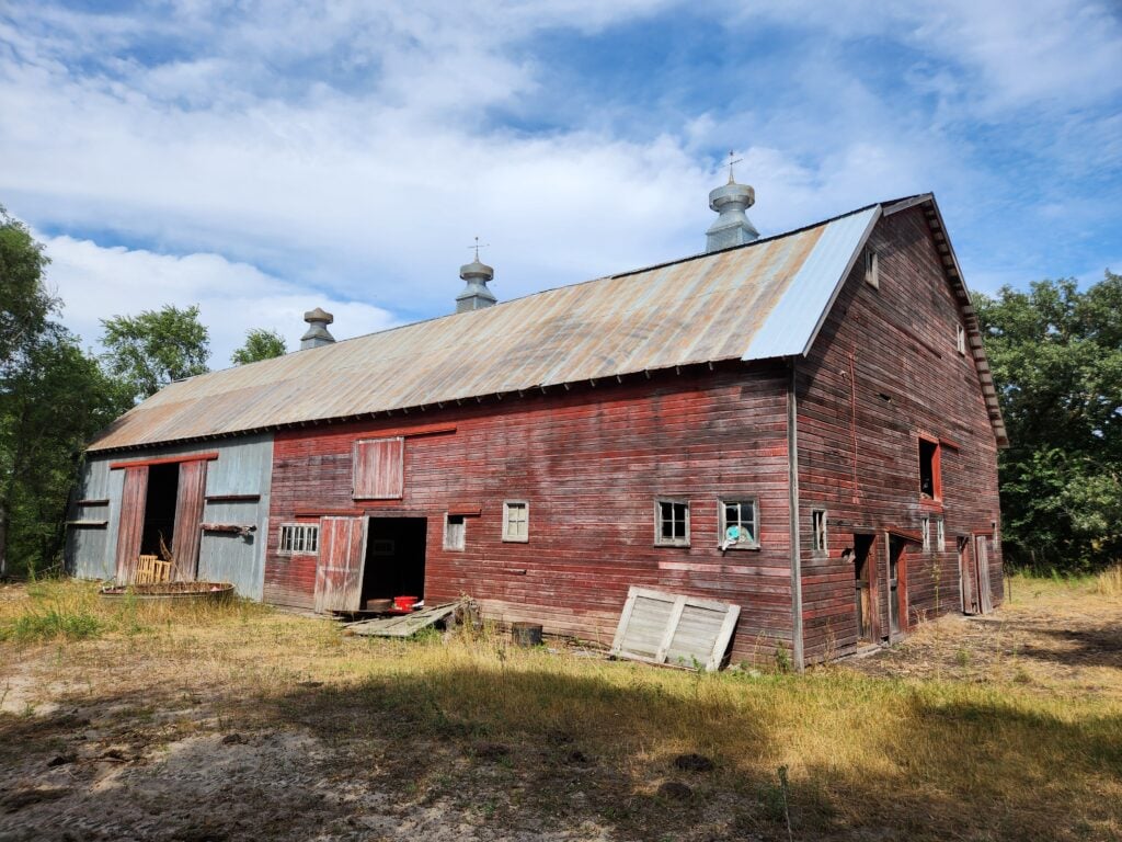 A large, old red wooden barn with a rusted metal roof stands on dry grass under a partly cloudy sky.