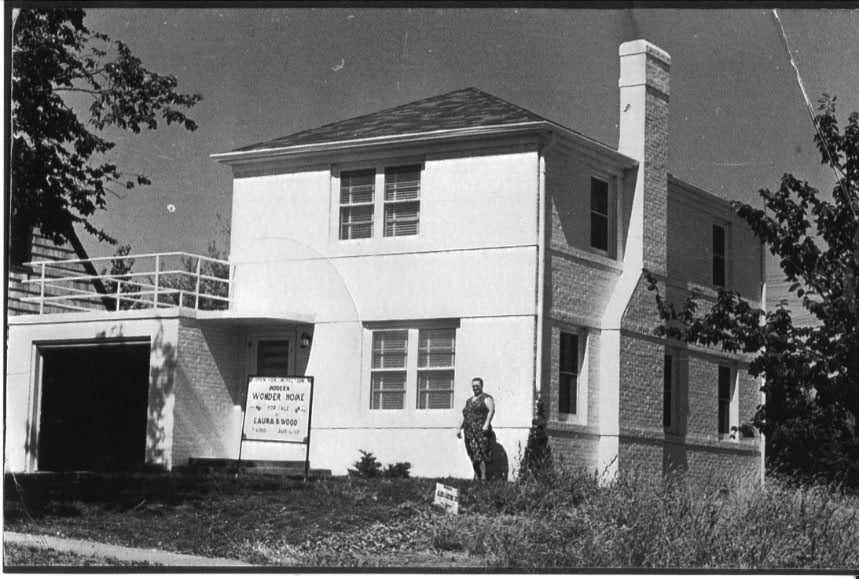 A person stands in front of a two-story house with a garage and a sign reading “Model House No. 14, Laura B. Swope, Owner.”.