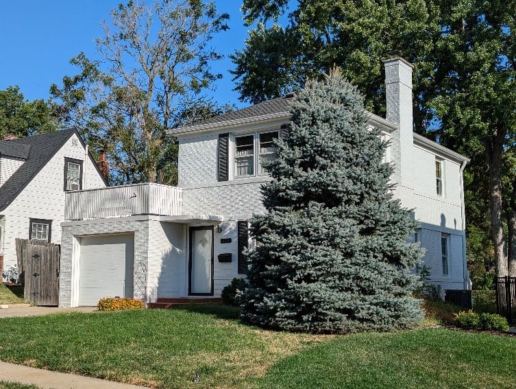 Two-story white brick house with attached garage, small balcony, and a large evergreen tree in the front yard. Neighboring house partially visible on the left.