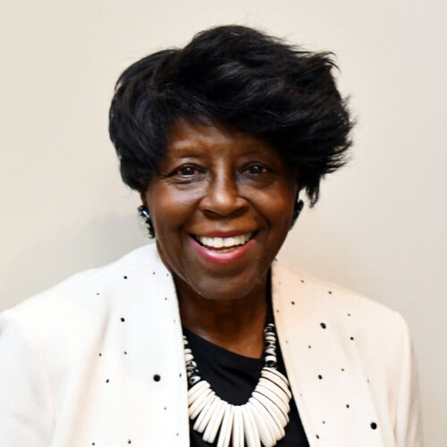 Smiling woman with short dark hair wearing a white jacket with black dots, a black top, and a chunky white necklace, standing in front of a plain light background.