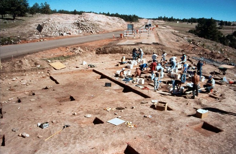 Archaeologists excavate a large site near a roadside, with equipment, tools, and multiple people working under clear skies.