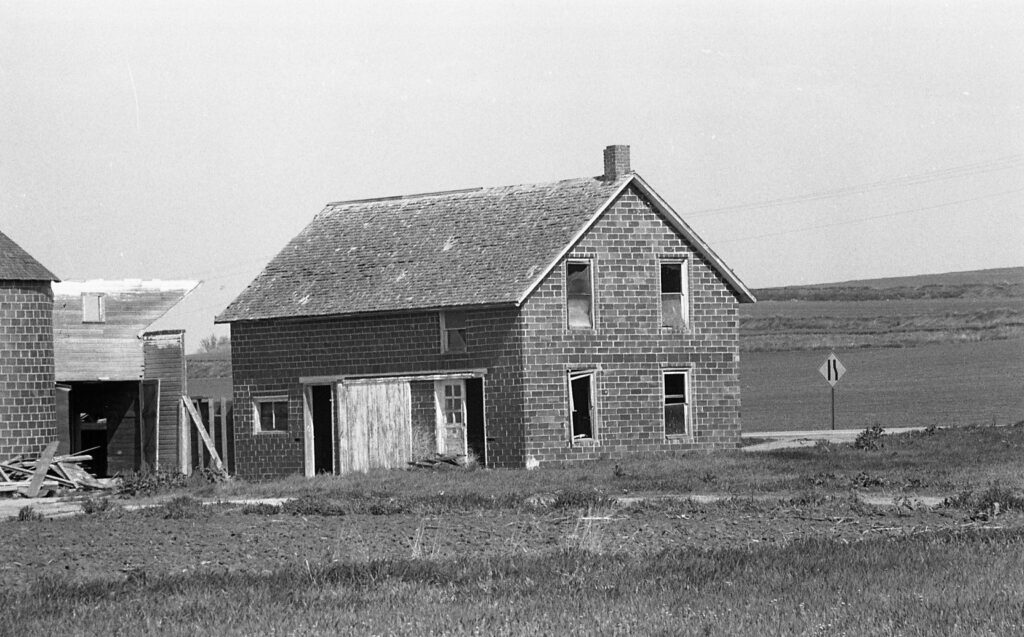 Black and white photo of an old, weathered two-story house with boarded windows and missing doors, situated near a rural road and open fields.