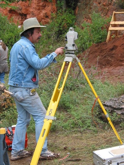 A person in a denim jacket and hat operates a surveying instrument mounted on a yellow tripod at an outdoor site with dirt and vegetation.
