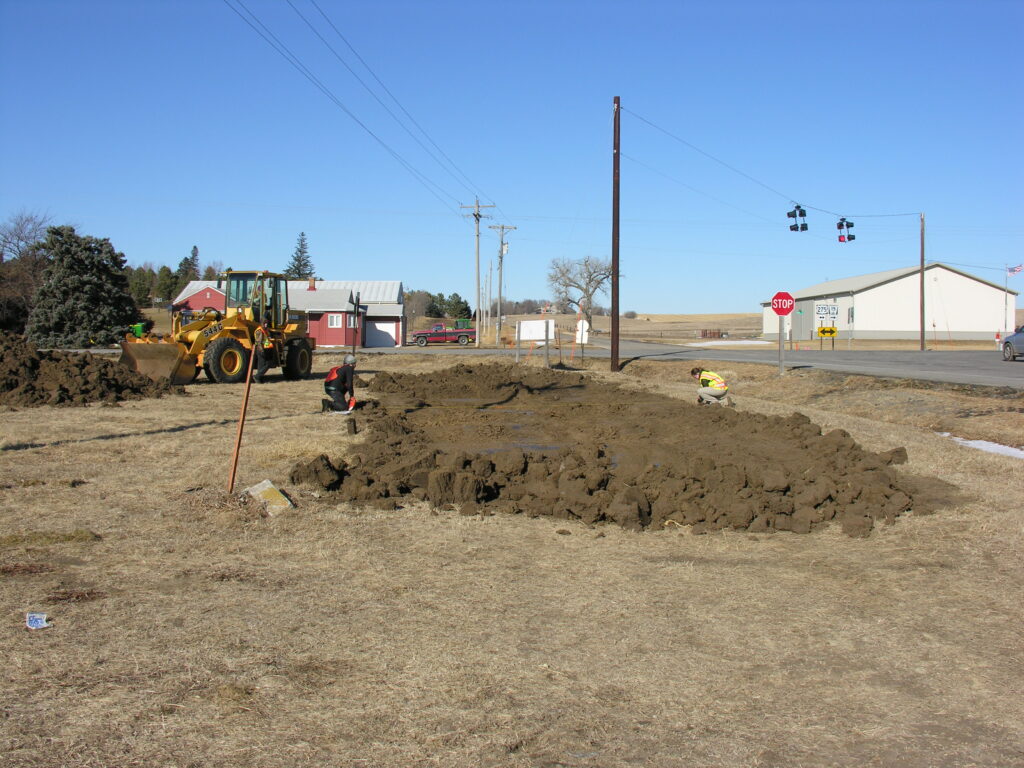 Two workers and a front-end loader are digging and moving soil near a rural intersection on a clear day.