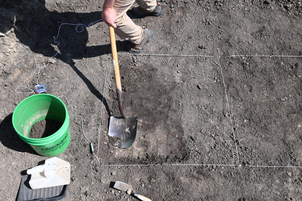 Person with a shovel stands near a green bucket and excavation tools, working within a marked-off rectangular area on dry soil.
