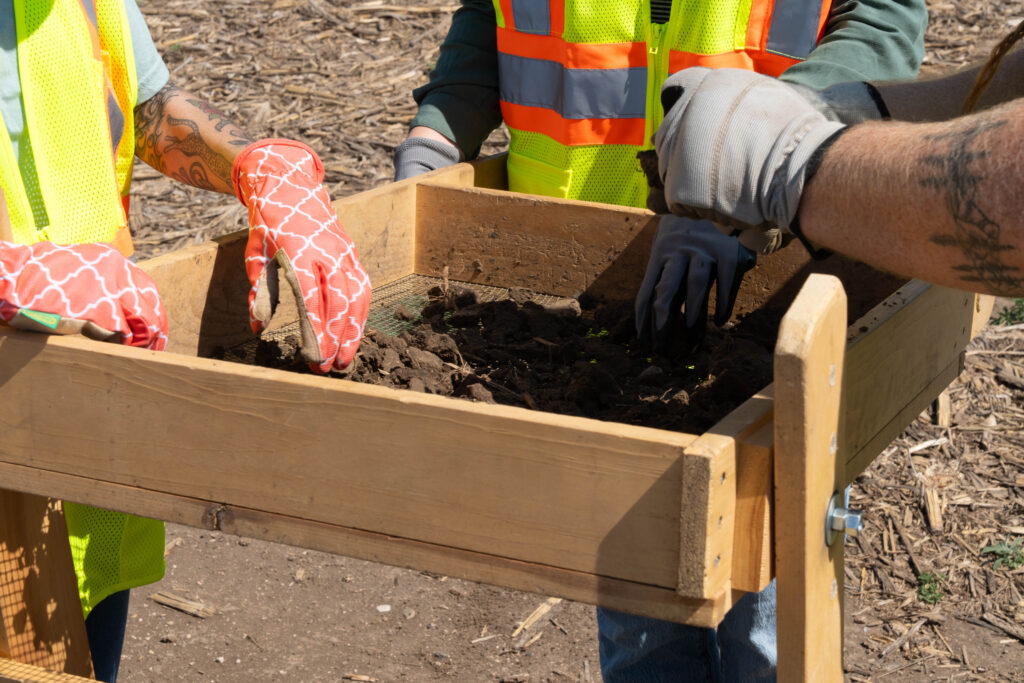 Three people wearing safety vests and gloves sift soil through a wooden frame outdoors, with dirt and debris visible on the screen.