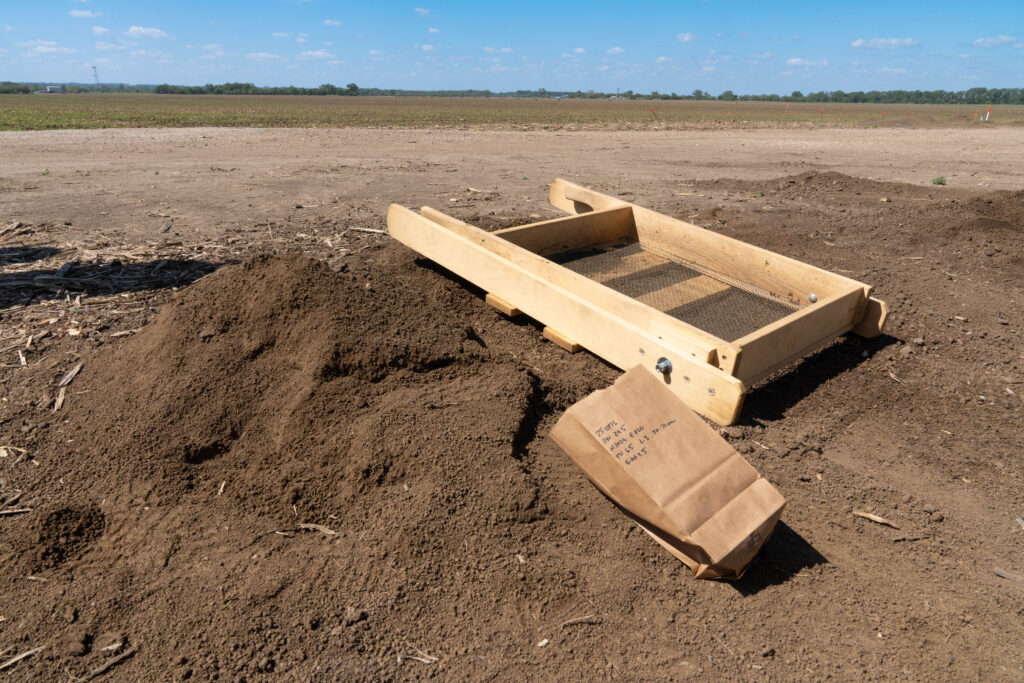 A wooden soil sifter and a brown paper sample bag rest beside a mound of dirt on an open, flat field under a blue sky.
