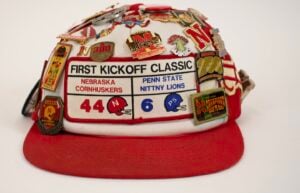 A red and white cap covered in pins, featuring a patch for the "First Kickoff Classic" football game between Nebraska Cornhuskers and Penn State Nittany Lions, score 44-6.