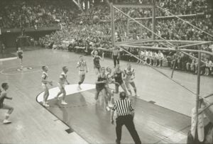 A black-and-white photo of a basketball game in progress, with players under the hoop and a referee watching, as a crowd fills the stands in the background.