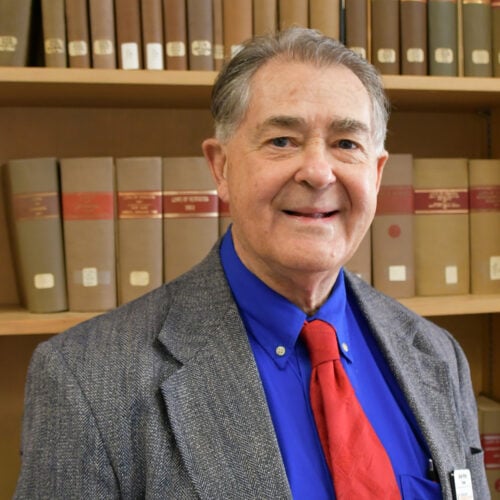 An older man in a gray blazer, blue shirt, and red tie stands in front of a bookshelf filled with large, labeled books.