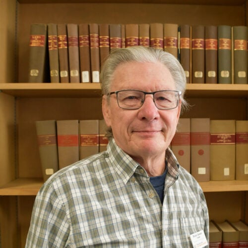 An older man with glasses and gray hair stands in front of a bookshelf filled with large books, wearing a plaid shirt and a name tag.