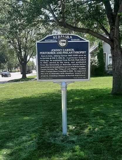 Nebraska historical marker sign for Johnny Carson, performer and philanthropist, stands on a grassy area near a sidewalk with trees and houses in the background.