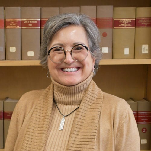Smiling woman with gray hair and glasses wearing a tan turtleneck sweater stands in front of shelves filled with books.