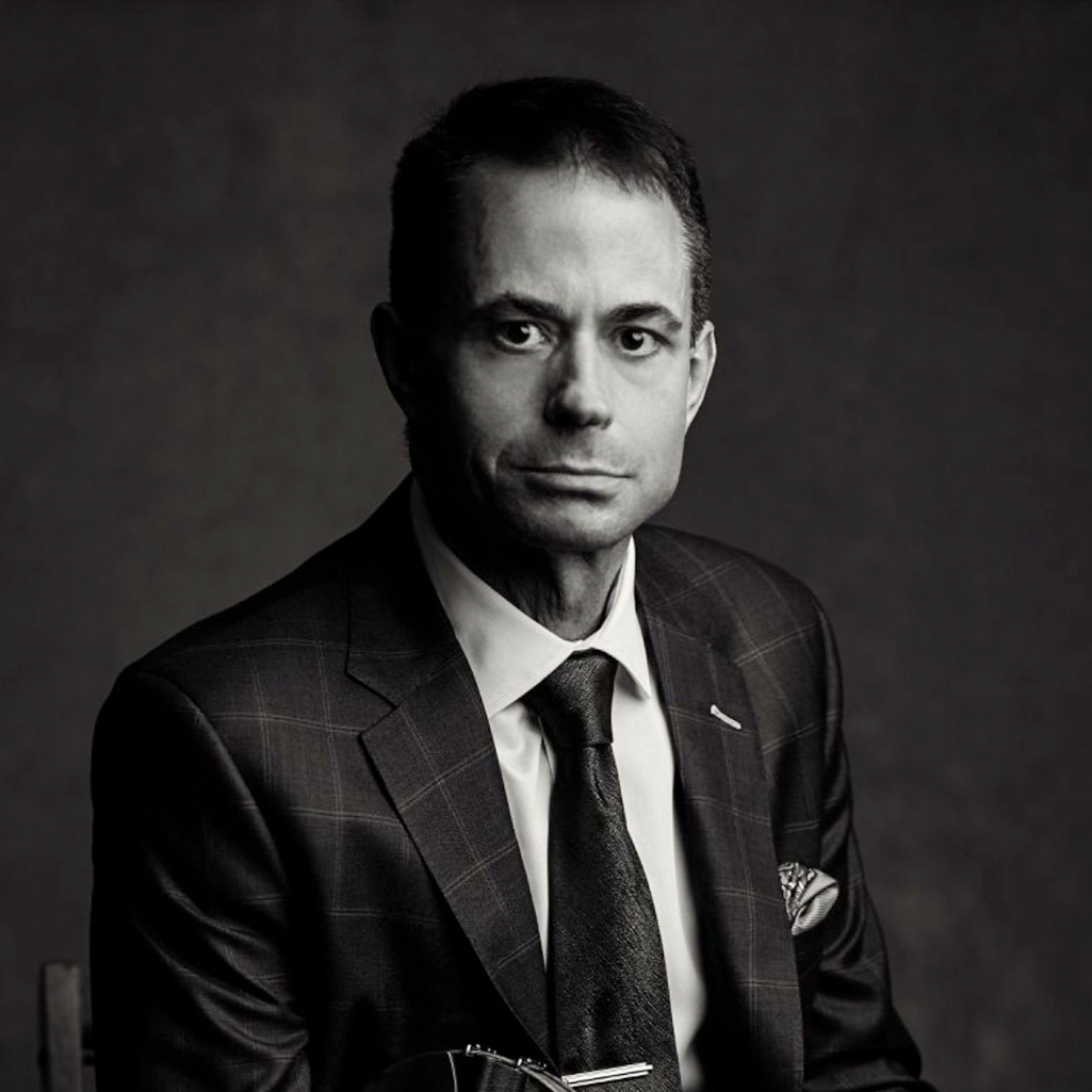 A man in a suit and tie sits for a formal portrait against a dark background, looking directly at the camera with a neutral expression.