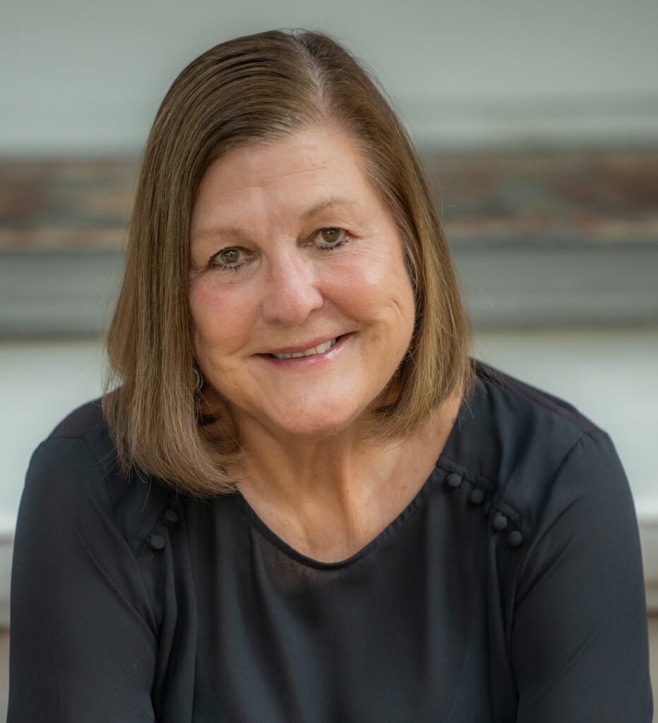 A middle-aged woman with straight, shoulder-length brown hair is smiling and wearing a black blouse, seated in front of a blurred background.