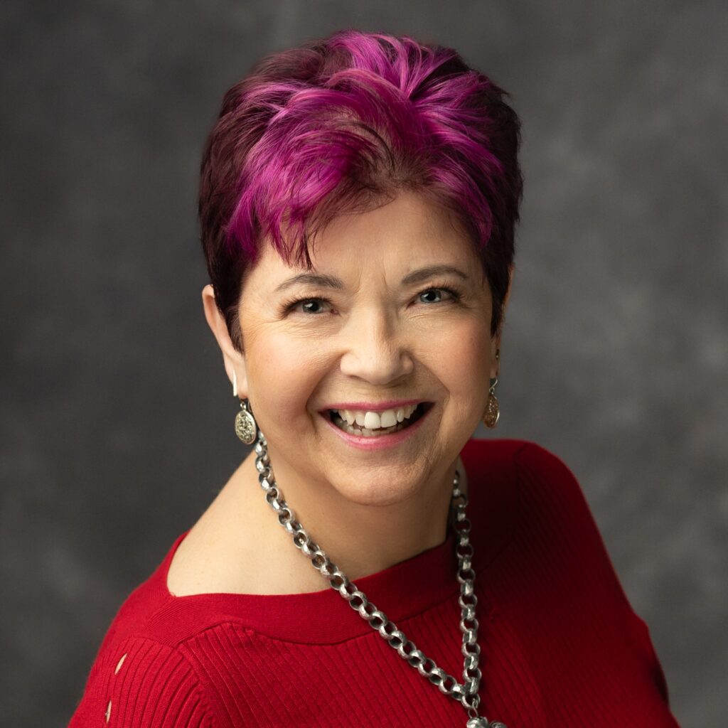Smiling woman with short purple and brown hair wearing a red top, silver chain necklace, and earrings, posed against a gray background.