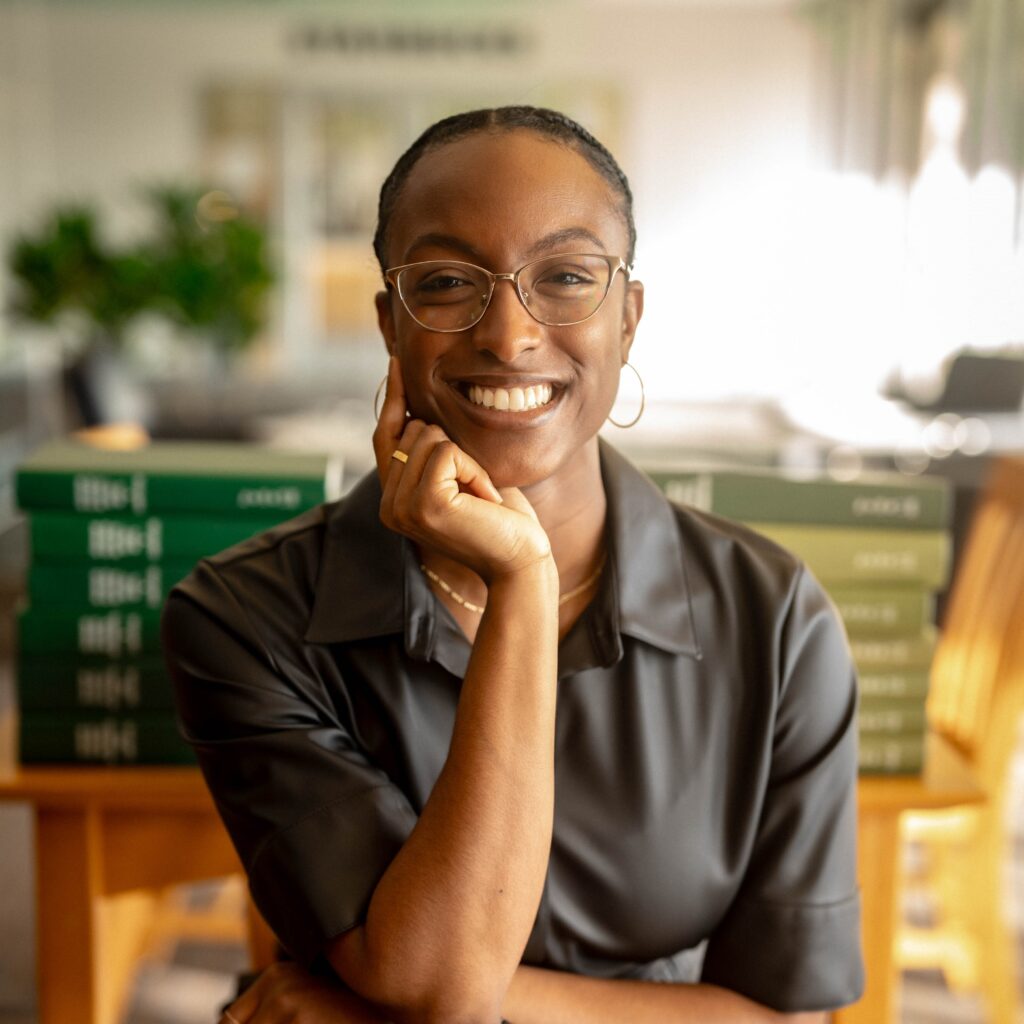 A person wearing glasses and a dark shirt smiles while resting their chin on their hand, with stacks of green books and a blurred background behind them.