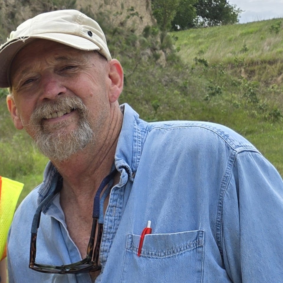 Man with a gray beard wearing a beige cap, blue shirt, and sunglasses around his neck standing outdoors in a grassy area.