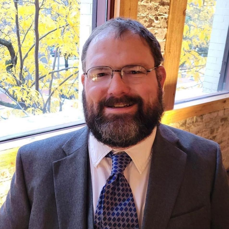 Man with glasses, a full beard, and a suit sits indoors by a window with fall foliage visible outside.