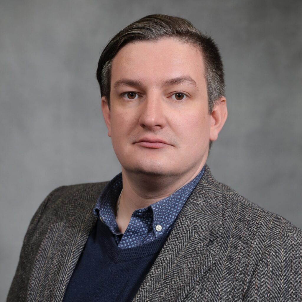 A man with short, dark hair wearing a gray patterned blazer, navy sweater, and blue polka dot shirt poses in front of a plain gray background.