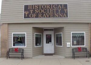 Street view of the Historical Society of Ravenna building with a white door, two windows, and benches decorated with red bows on either side of the entrance.