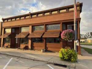 A red brick building with large windows and brown awnings, flower planters by the entrance, and a street visible in the foreground.