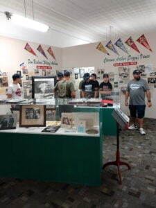 A group of people view baseball memorabilia displayed in glass cases at a museum, with Hall of Fame plaques and photos on the walls.