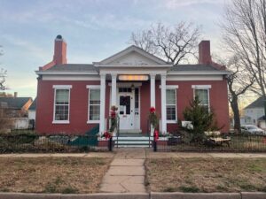 A red brick house with white pillars, decorated with holiday wreaths and bows, sits behind a black iron fence on a quiet residential street.
