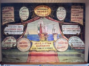 A vintage illustrated advertisement features a ship at sea, surrounded by oval-shaped ads for various local businesses, including hardware, groceries, lumber, and dry goods.