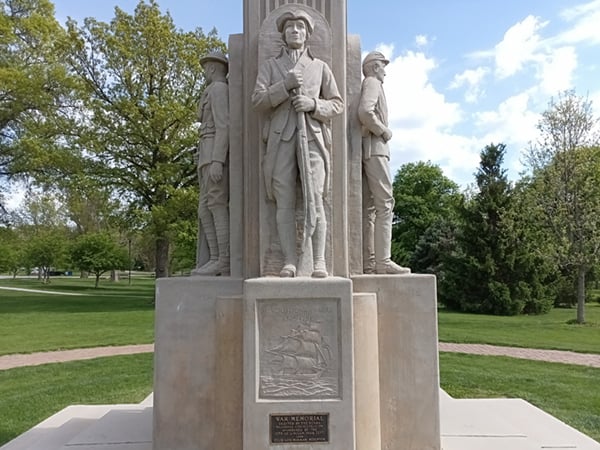 Three stone statues of soldiers stand on a pedestal in a park, with a plaque and engraved image of a ship on the front of the monument. Trees and grass are visible in the background.