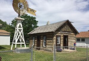 A small log cabin labeled "U.S. Post Office, Fairview, MT 59221" stands beside a tall white windmill, surrounded by a chain-link fence and neighboring buildings.
