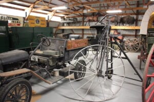 Antique vehicles and machinery are displayed in a museum, including a vintage truck with a "Museum" sign, wagons, and animal taxidermy on a shelf in the background.