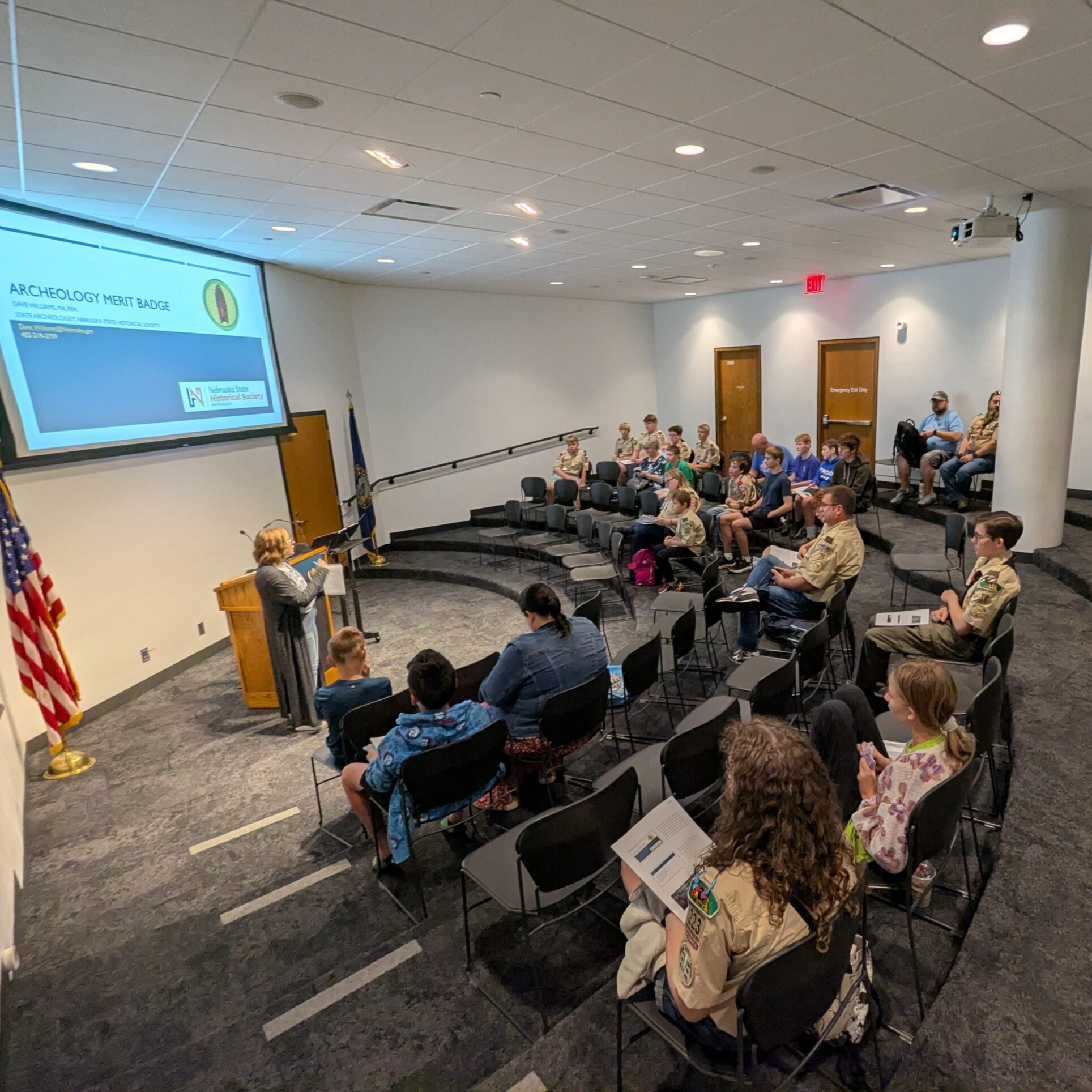 A group of people, including scouts in uniform, sit in a lecture hall listening to a speaker at a podium presenting about the Archeology Merit Badge.
