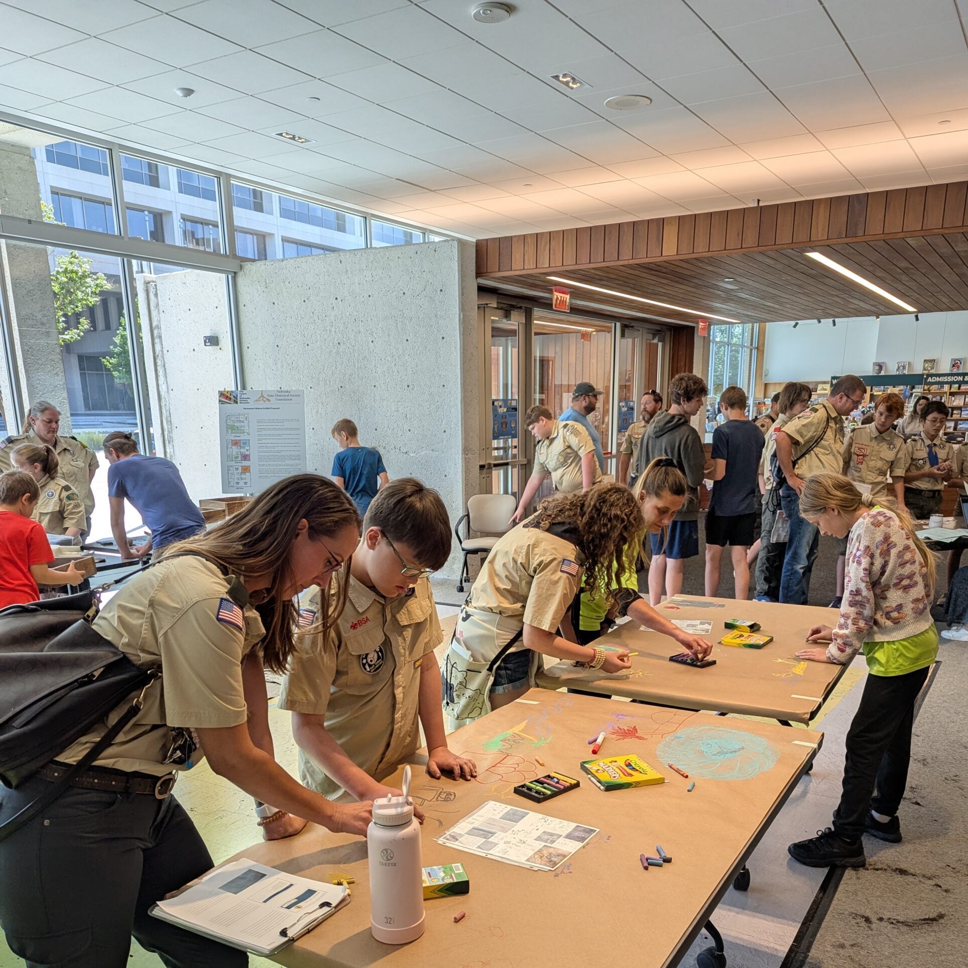 A group of scouts and other participants work on crafts and activities at tables in a busy, well-lit indoor space during Summer Workshops.
