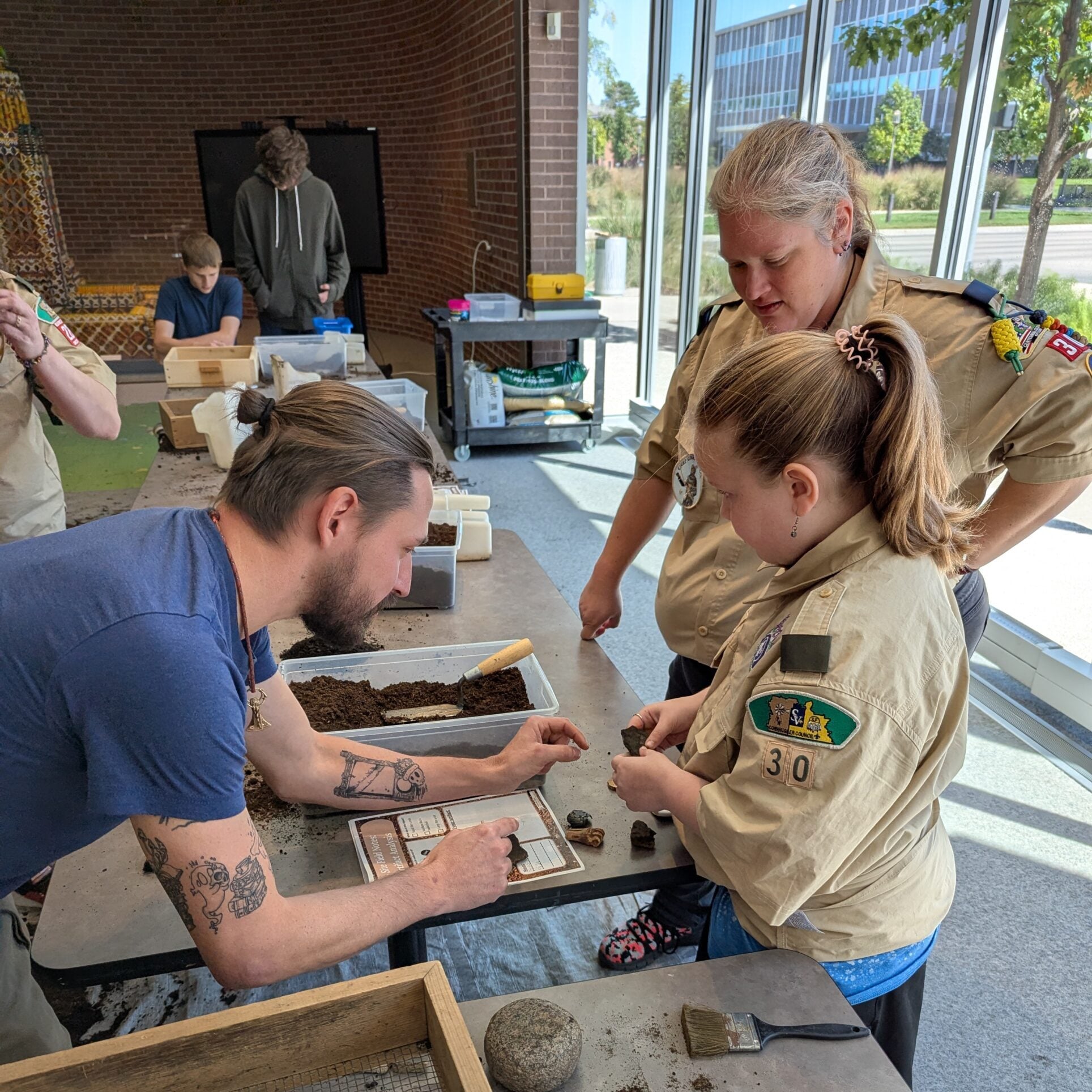 A man leads two people in scout uniforms through hands-on excavation with tools and soil samples indoors near large windows, as part of engaging Summer Workshops.