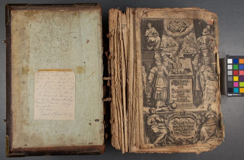 An old, worn book displayed open; left side shows handwritten note, right side features intricate illustrated title page with figures and decorative elements. Color calibration card beside book.