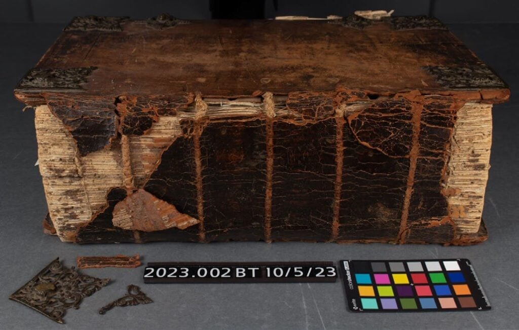 Large, old book with damaged leather cover, exposed spine, and missing corners. Metal fittings are detached and placed nearby. A color calibration chart and ID label are included.